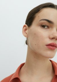 Nahaufnahme des Gesichts eines Modells, mit glatter Haut und Unreinheiten, braunen Augen und vollen Lippen. Das Modell trägt ein rot-orangefarbenes Hemd mit Kragen.