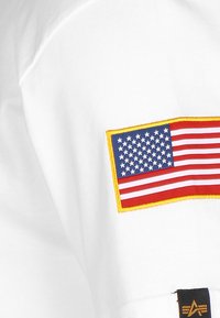 Hvit T-skjorte med et brodert amerikansk flaggmerke i rødt, hvitt og blått, omgitt av gult. Laget av myk bomull.