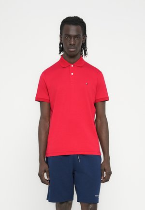 Muškarac stoji na bijeloj pozadini, nosi crvenu polo majicu kratkih rukava i tamnoplave kratke hlače, gleda naprijed s opuštenim rukama.