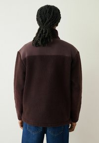 Burgundy fleece jacket with a high collar, featuring a smooth upper section and ribbed accents on the sleeves. Textured, warm, and soft.