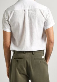 Camicia bianca di lino a maniche corte con cucitura a yoke sul retro, abbinata a pantaloni verde oliva con tasche posteriori con bottone e dettaglio del tag.