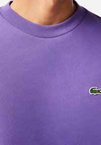 Sudadera de algodón púrpura con un cuello acanalado y un pequeño logo bordado de cocodrilo en verde y rojo en el frente.
