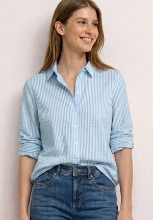Frau lächelt, trägt ein hellblau-weiß gestreiftes Hemd mit hochgekrempelten Ärmeln, in mittelblau gewaschene Jeans gesteckt, vor einfarbigem Hintergrund.