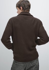 Brązowy sweter prążkowany z szerokim kołnierzem i teksturowanym wykończeniem. Tył ma dopasowaną talię oraz subtelne szwy, zestawiony z szarymi spodniami.