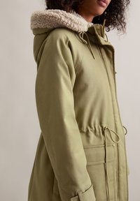 Olivengroene parka met een zachte beige capuchonrand, taillekoord en voorzakken, met een gestructureerde buitenstof en een veilige ritsluiting.