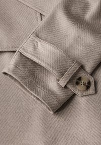 Beżowa tkanina kurtki w wzór jodełki, z złożonym mankietem i detalem guzika, ukazująca miękką teksturę i precyzyjne szycie.