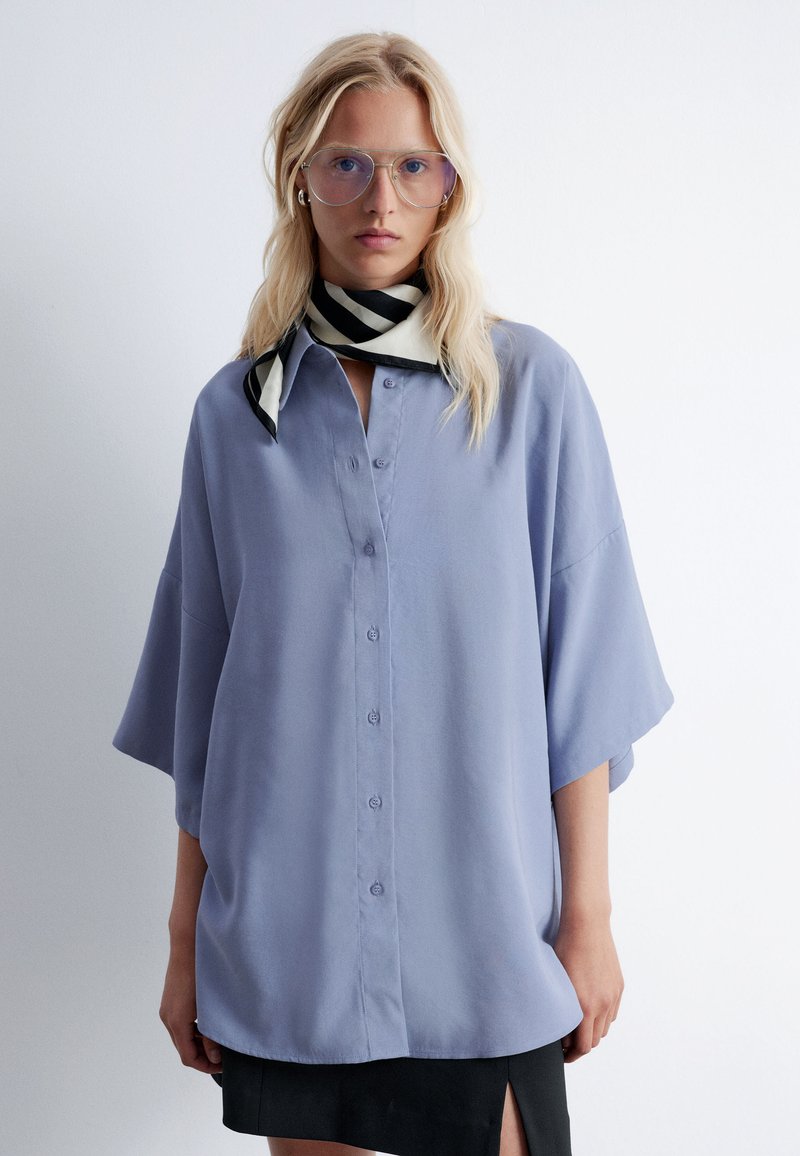 Light purple, loose-fitting blouse with a button-down front, short sleeves, and a smooth texture. Accented with a black-and-white striped scarf.