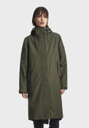Femme portant un long imperméable à capuche vert olive avec des poches frontales et des fermetures à boutons-pression, debout devant un fond uni.