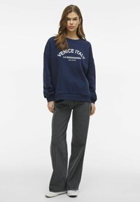 Mornarska dukserica s bijelim grafičkim printom "Venecija Italija"; opušteni kroj i okrugli izrez. Uparena s tamnim trapericama visokog struka i bijelim tenisicama.