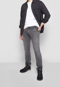Homme portant un jean gris, des baskets noires montantes, un t-shirt blanc et une chemise noire déboutonnée avec les manches retroussées, debout devant un fond uni.