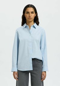 Camicia azzurra chiara button-up realizzata in tessuto morbido, con colletto classico e maniche lunghe con polsini abbottonati, indossata con pantaloni grigi.