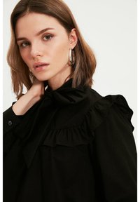Blusa nera con colletto e polsini arricciati, chiusura con bottoni e texture liscia. Il modello indossa grandi orecchini a cerchio. Illuminazione soffusa.