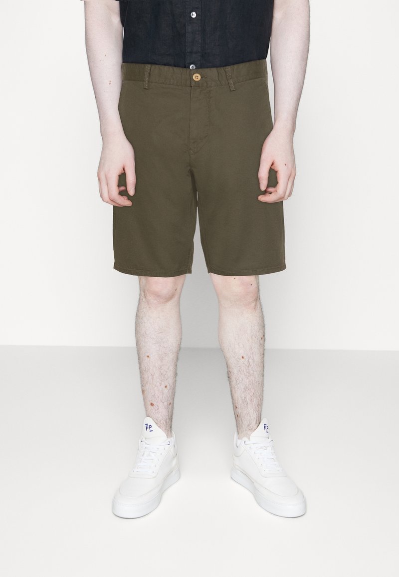 Personne portant un short olive mi-long, des baskets blanches et une chemise noire partiellement visible, debout sur un fond clair.