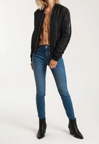 Černá bomber bunda s žebrovanými manžetami, spárovaná s hnědým zapínacím kardigánem a modrými skinny džíny. Dokončeno černými kotníkovými botami.
