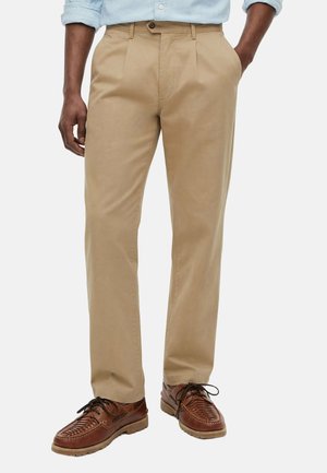 Mann trägt beige gerade geschnittene Hose mit Seitentaschen und braune geflochtene Lederschnürschuhe, Hände in den Taschen.
