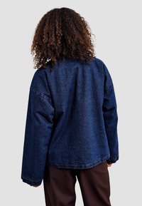 Tamno plavi traper jakna s opuštenim krojem, dugim rukavima i blago zakrivljenim rubom. Suptilni šavovi duž rubova.