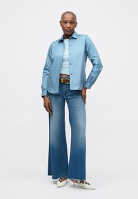 Donna che indossa una camicia leggera blu sopra una maglietta bianca, jeans larghi blu con orli sfilacciati, cintura marrone e tacchi bianchi, in piedi con sicurezza.