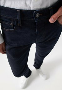 Jeans de mezclilla azul oscuro con costuras naranjas, diseño de cinco bolsillos y cierre de botón. Llevados con zapatillas blancas y una camisa clara.