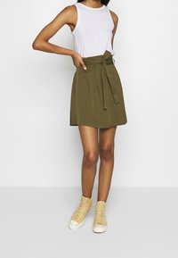 Olivengrünen Rock mit einem Bindegürtel, Falten und einer lockeren Passform, kombiniert mit einem weißen ärmellosen Oberteil und beigen High-Top-Sneakern.