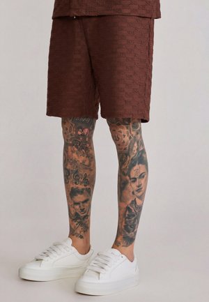 Dolne partie nóg z detalicznymi czarno-szarymi tatuażami twarzy, nut muzycznych oraz wzorów, ubranych w brązowe teksturowane spodenki i białe trampki.