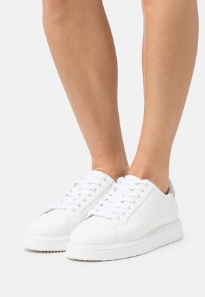 Person wearing clean white leather lace-up sneakers with thick soles, standing on a plain light background.