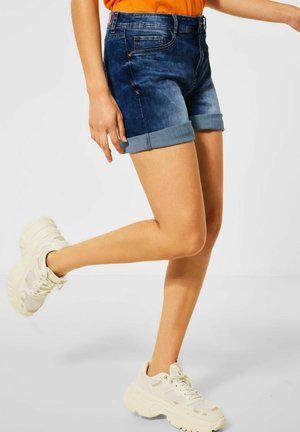 Person trägt dunkelblaue aufgekrempelte Jeansshorts, ein orangefarbenes Top und klobige weiße Sneakers und steht auf einem Bein vor einem einfarbigen Hintergrund.