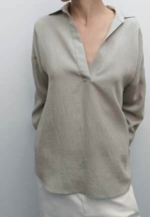 Personne portant un haut ample à manches longues beige avec un col en V profond, associé à un pantalon de couleur claire, sur un fond uni.