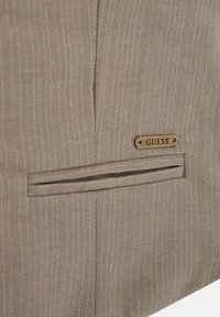 Smeđa tkanina s prugicama koja sadrži malu, uvučenu mesinganu pločicu s oznakom "GUESS", s horizontalnim detaljem džepa sa prorezom.