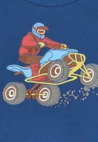 Pilota con casco e attrezzatura rossa salta su un quad blu con telaio giallo, sollevando polvere su uno sfondo di tessuto blu.