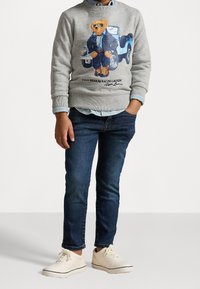 Grå sweatshirt med en björngrafik i en blå outfit, blå denimjeans och ljusa skor med snörning; lager på lager med en blå skjorta under.