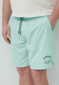 Muntgroene zwembroek met een elastische tailleband en een trekkoord. Heeft een geprint ontwerp met tekst en een haai-illustratie op het been.