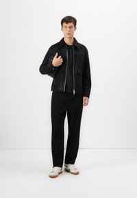 Jonge man in zwarte jas, zwarte broek, witte sneakers, met een zwarte clutch tas in zijn handen, staande tegen een plain witte achtergrond.