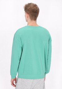Mentazöld pulóver hosszú ujjakkal és kerek nyakkal, sima textúrával, bordázott mandzsettákkal és alsó résszel. Szürke melegítőnadrágon viselve.