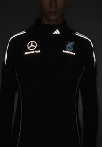 Sort langærmet jakke med lynlås, med reflekterende detaljer og logoer for Mercedes-AMG og Petronas på brystet og ærmerne.
