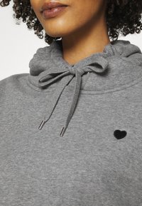 Felpa grigia con cappuccio regolabile, piccola ricamo a cuore nero e accessori in metallo argentato sui lacci. Tessuto morbido.