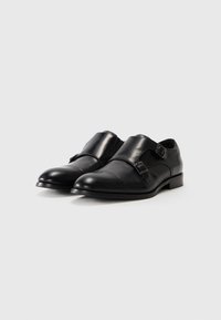 Scarpe in pelle nera con una finitura lucida, caratterizzate da un design a doppia fibbia, punta arrotondata e dettagli di cucitura minimi. Tacco basso.