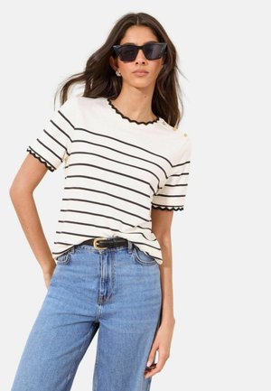 Woman wearing black cat-eye sunglasses, white short-sleeve shirt with black horizontal stripes and scalloped edges, tucked into blue jeans with black belt.