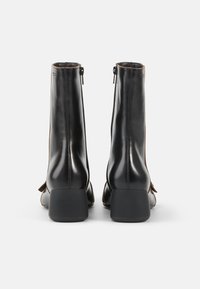 Botas de tobillo de cuero negro con textura suave, tacones cuadrados, cierre de cremallera y diseño minimalista, vista trasera que muestra el detalle de la costura.