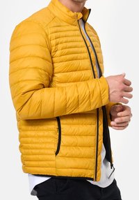 Gelbe gesteppte Jacke mit Stehkragen, Reißverschluss vorne und schwarzen Akzenten, mit horizontalen Nähten und seitlichen Taschen.