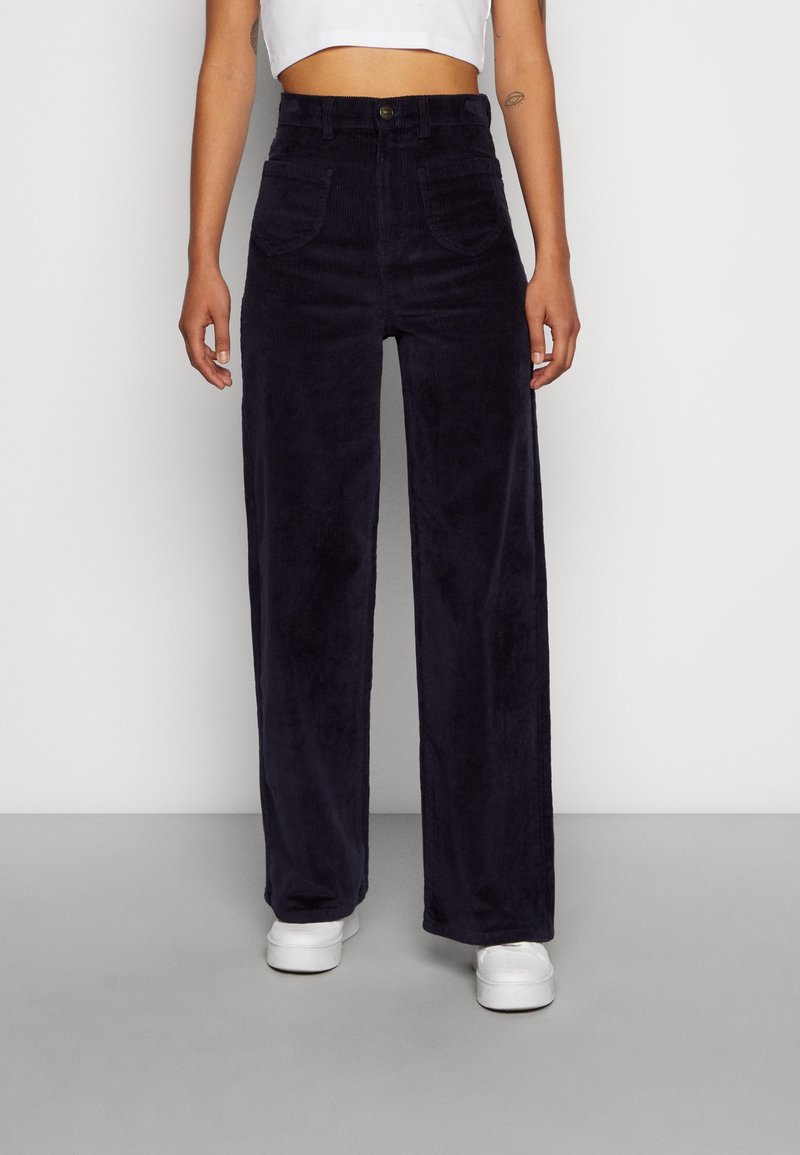 Personne portant un pantalon noir en velours côtelé taille haute à jambes larges et des baskets blanches, debout sur un sol gris devant un mur blanc.