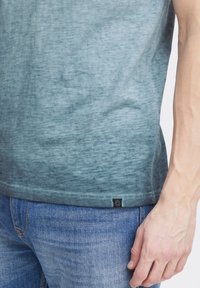 Camiseta de gradiente verde, de manga corta, hecha de tela suave. Presenta un dobladillo redondeado y una pequeña etiqueta de marca cerca de la parte inferior.