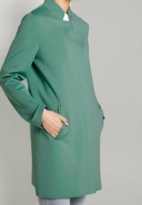 Groene jas met een hoge kraag, lange mouwen en zijzakken. Gladde textuur en minimale naden, voor een getailleerd silhouet.