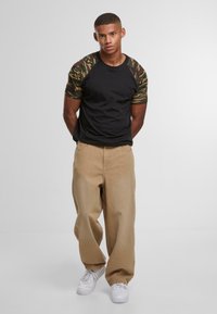 Sort kortærmet skjorte med camouflage-mønstrede ærmer, parret med beige vide bukser og hvide sneakers. Glat tekstur hele vejen igennem.