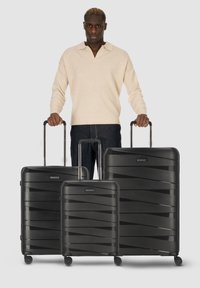 Trois valises rigides noires de différentes tailles présentent un design texturé et ondulé, avec des poignées rétractables et des roues pour un transport facile.