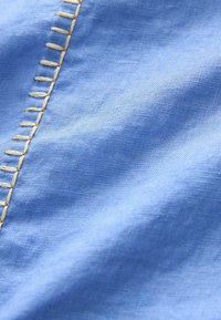 Tissu bleu avec broderie bouclée beige cousue main le long du bord, montrant la texture et des plis délicats.