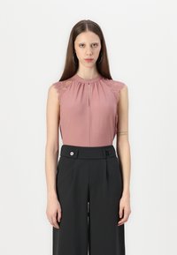 Haut sans manches en rose poussiéreux avec des accents en dentelle sur les épaules, présentant un col plissé et une ouverture en forme de clé. Associé à un pantalon noir ajusté.