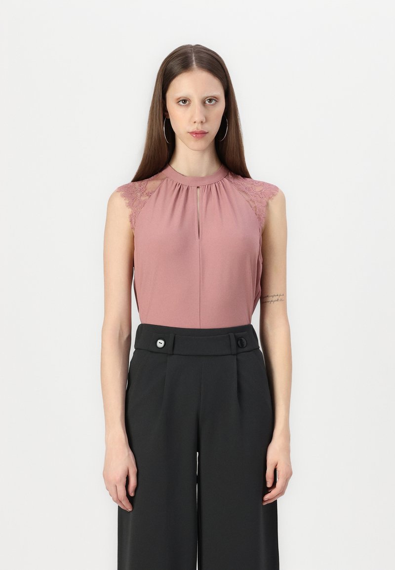 Haut sans manches en rose poussiéreux avec des accents en dentelle sur les épaules, présentant un col plissé et une ouverture en forme de clé. Associé à un pantalon noir ajusté.