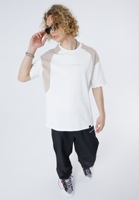 White t-shirt with beige raglan sleeves, featuring subtle logo print. Paired with loose black pants and gray sneakers.