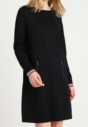 Kvinde iført en sort langærmet kjole med manchetter med røde, hvide og sorte striber og to lodrette lynlåslommer foran.