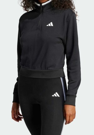 Woman wearing black long-sleeve athletic top with half-zip and matching black leggings, both with white Adidas logos.